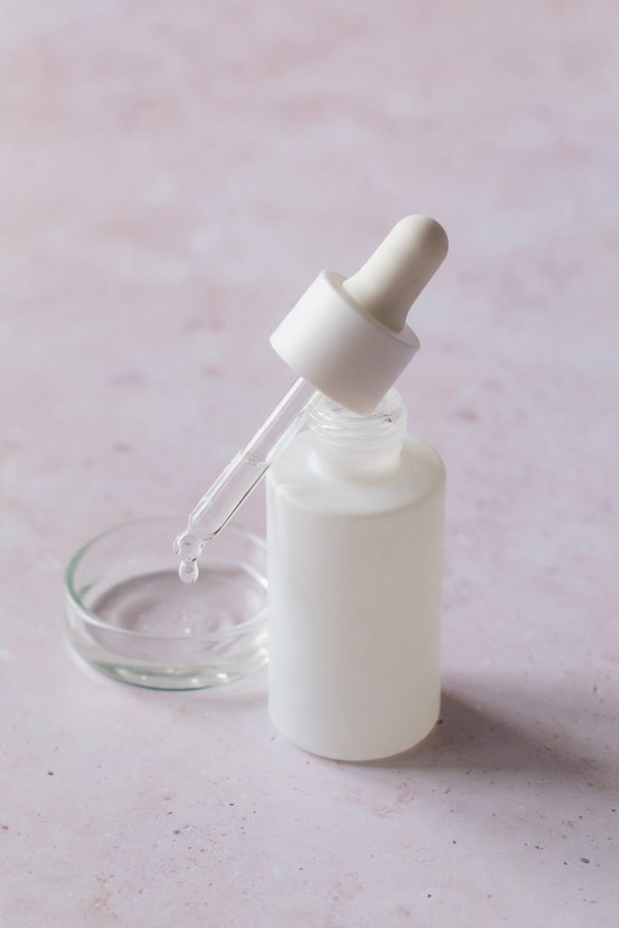 Frosted dropper serum bottle with a drop of serum beside a small glass dish